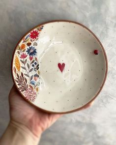 a hand holding a small bowl with flowers and hearts painted on the side, in front of a white background