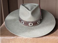 The Bandit is a stand out hat that captures the feeling of the American West. . The base of the hatband is distressed leather that is then layered with imprinted brown leather, bolo sliders and lots of nailspots. It is finished with this colorful feather combo. The brim of the LID custom hat body is 4" in diameter and the crown is 4.5". This is a stiff hat. Available in Putty or Pecan All hats are made to order. Allow up to 4 weeks for your hat to ship. Western Style Brown Hat With Flat Crown, Adjustable Distressed Hats For Country Events, Distressed Adjustable Hats For Country Events, Flat Brim Hats With Leather Sweatband For Country Events, Brown Felt Hat With Flat Crown For Country Events, Brimmed Hats With Leather Sweatband For Country Events, Brown Flat Crown Felt Hat For Country Events, Brown Concho Hat For Rodeo, Rustic Brown Hat Bands For Rodeo