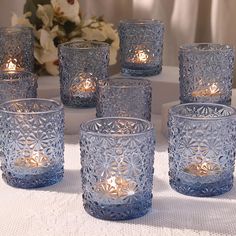 blue glass candles are sitting on a white tablecloth with flowers in the back ground