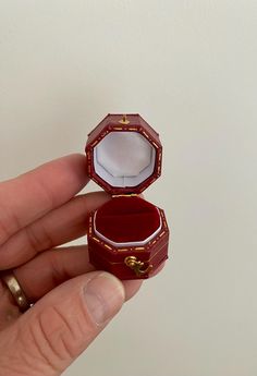 a hand holding an open red ring box with gold trimmings on the inside