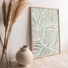 a white vase sitting next to a plant and a framed print on the wall behind it