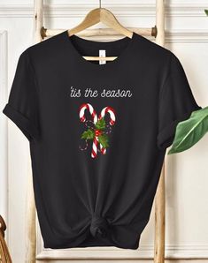 🎄Celebrate the holidays in style with this Tis The Season T-Shirt! Featuring two classic candy canes crossed together with a sprig of holly and berries, accompanied by the festive phrase 'Tis the Season in elegant white script font, this shirt is the perfect blend of sophistication and holiday cheer. 🍬✨ Ideal for Christmas parties, family gatherings, or cozy holiday shopping trips, this stylish t-shirt adds a touch of festive charm to your wardrobe. The design captures the essence of the seaso Black Christmas T-shirt As Gift, Black Crew Neck Top For New Year, Black Crew Neck T-shirt For Holidays, Black Christmas Graphic Tee, New Year Holiday Crew Neck T-shirt, Black Holiday Tops With Letter Print, Black Holiday Top With Letter Print, Black Casual T-shirt For Festive Occasions, Casual Black T-shirt For Festive Occasions