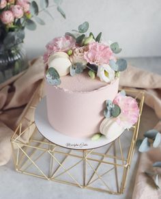 a pink and white cake with flowers on top sitting on a gold wire stand next to a potted plant