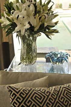 flowers in a vase on a glass table with two pillows and a window behind it