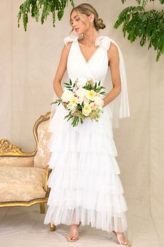 a woman in a white dress holding a bouquet and standing next to a gold chair