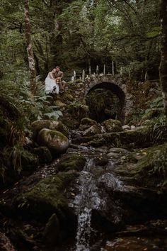 a bride and groom are sitting on the mossy rocks in front of a bridge