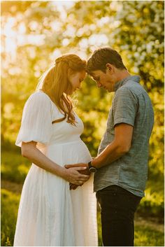 a pregnant couple standing next to each other in front of trees and grass at sunset