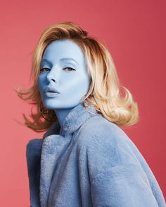 a woman with blue paint on her face and hair is standing in front of a pink background