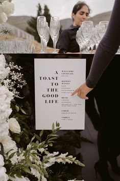 a woman pointing at a sign that says toast to the good life with wine glasses