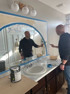 two men standing in front of a bathroom mirror with blue tape on the edge of it