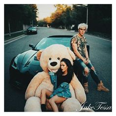 two people sitting next to a large teddy bear on the side of the road with a car behind them