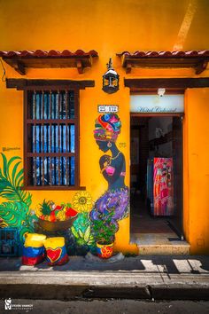 a yellow building with potted plants in front of it and a painting on the wall