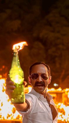 a man holding up a beer bottle with flames in the background