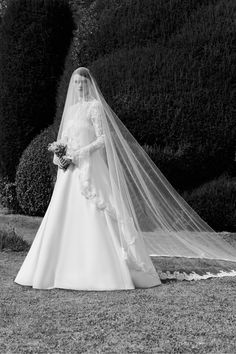 a woman in a wedding dress and veil is standing on the grass near some bushes