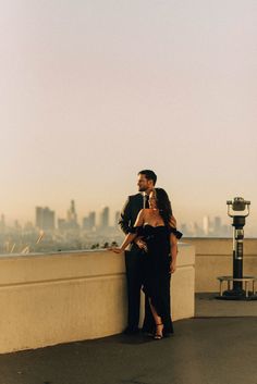 a man and woman standing next to each other on top of a roof with the city in the background