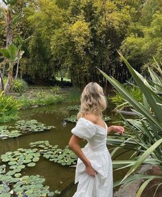 a woman in a white dress standing next to a pond with lily pads on it