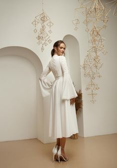 a woman standing in front of a white wall wearing high heels and a dress with long sleeves