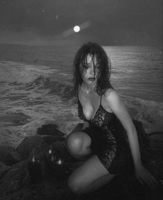 black and white photograph of a woman sitting on rocks near the ocean at night with full moon in background