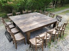 a large wooden table surrounded by chairs in the middle of a gravel area with trees behind it