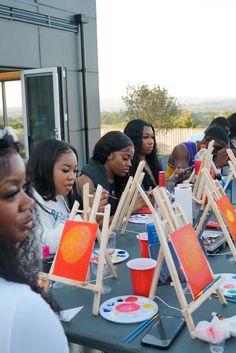 a group of women sitting around a table with paintings on easels in front of them