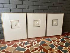 three framed pictures are on the floor in front of a brick wall and tiled floor