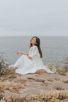 a woman sitting on top of a sandy beach next to the ocean wearing a white dress