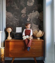 a young boy sitting on top of a dresser next to a wallpapered wall