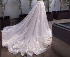 the back of a wedding dress with white flowers on it, in front of some potted plants