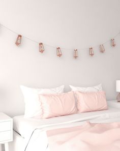 a white bed topped with pink pillows next to a night stand and lamp on the wall