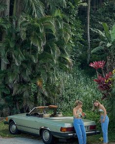 two women standing next to a green car in front of some palm trees and flowers