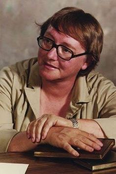 a woman wearing glasses sitting at a table with a book in front of her and looking off to the side