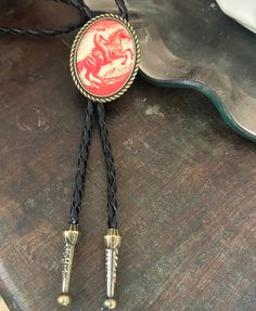 This awesome bolo tie has a vintage cowboy print with a clear resin coating and metal backing. Handmade in our shop! The cord is black and genuine leather. Our bolos pair nicely with many of our belt buckles! They make wonderful gifts. The western bolo tie rope length is 39' ; pendant size is 1 1/2'' x 1 1/4'' Handmade Adjustable Bolo Tie For Rodeo, Handmade Adjustable Bolo Ties For Rodeo, Western Bolo Ties For Western-themed Events, Western Style Adjustable Bolo Ties, Western Bolo Ties With Adjustable Length For Western-themed Events, Western Bolo Ties With Adjustable Length, Vintage Adjustable Hand Tooled Bolo Tie, Adjustable Vintage Hand Tooled Bolo Tie, Hand Tooled Western Bolo Ties As Gift