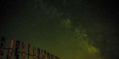 the night sky is filled with stars above a wooden fence and some green lights in the background