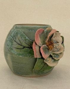 a green vase with a flower on the top and leaves on the bottom, sitting on a white surface