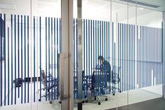 a man sitting at a desk in front of a glass wall with blue vertical blinds