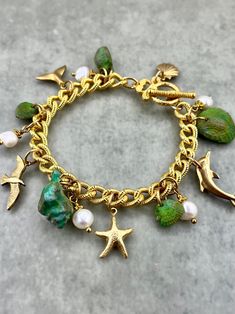 WHAT IT IS: A "Seaside Soirée" theme charm bracelet featuring hand-painted vintage lucite seashells, vintage solid brass charms, and freshwater pearls, all dangling from a 10k gold plated chain with a toggle clasp.  THE SPECS: - 7.5 inches total length. - Toggle clasp closure. WHY IT'S SPECIAL: The vintage lucite seashells began in subtle hues of white, tan, and coral. While lovely as they were, we meticulously hand-painted them in various layers to add depth and playful, colorful charm. With the transformed, vibrant shells hung from an elegant gold-plated chain and adorned with  genuine freshwater pearls, this "Seaside Soirée" bracelet balances colorful whimsy with tasteful sophistication--capturing the lively essence of a coastal celebration. This bracelet comes in purple too! Take a loo Bohemian Style Bracelet With Starfish Charm, Artistic Adjustable Jewelry For Beach, Beach Charms Bracelet Jewelry, Turquoise Ocean-inspired Bracelet With Lobster Clasp, Ocean-inspired Charm Bracelet As A Gift, Handmade Dangle Bracelets For Beach, Ocean-inspired Charm Bracelet Gift, Beach Ocean-inspired Charm Bracelet, Handmade Charm Bangle For Beach