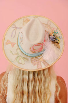 Add some whimsy to your wardrobe with THE CHARLOTTE RANCHER HAT. This cream hat features playful gold accents, pastel painted bows, and a charming blue ribbon band. Adorned with pink and gold feathers and a delightful floral combo, this hat is perfect for those who love a good bow.