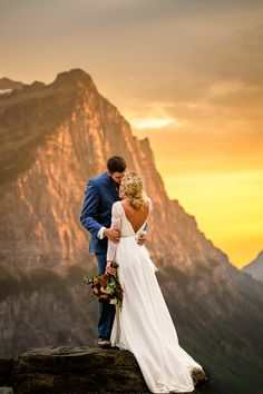 a bride and groom standing on top of a mountain with the sun setting behind them