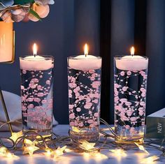 three lit candles with pink flowers in them on a table next to a flower arrangement