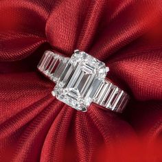 an emerald - cut diamond ring sits atop a red satin fabric