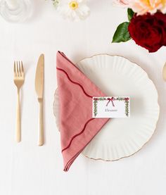 a place setting with pink napkin, silverware and flowers on the white tablecloth