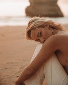 a woman sitting on top of a sandy beach next to the ocean with her head down