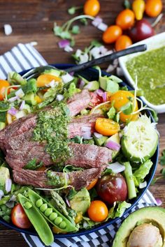 a salad with meat and veggies in it on a table next to an avocado