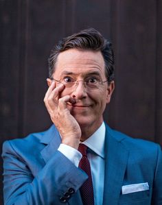 a man in a blue suit and tie with his hand on his chin looking at the camera