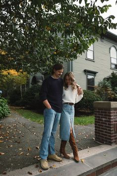 a man and woman standing on the sidewalk in front of a house with leaves all around them