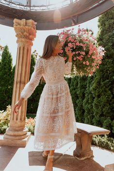 a woman in a white dress standing next to a stone pillar and flower pot with pink flowers on it