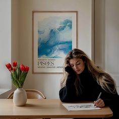 a woman sitting at a table writing on a piece of paper next to a vase with red tulips