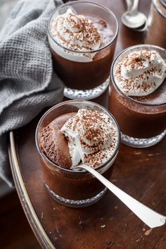 three glasses filled with dessert sitting on top of a wooden table next to spoons