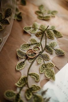 two wedding rings on top of a table with green leaves and lace around it, next to a piece of paper