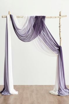 a purple and white wedding arch with sheer drapes on the top, hanging from a wooden pole